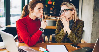 Eine Frau flüstert ihrer Arbeitskollegin etwas zu